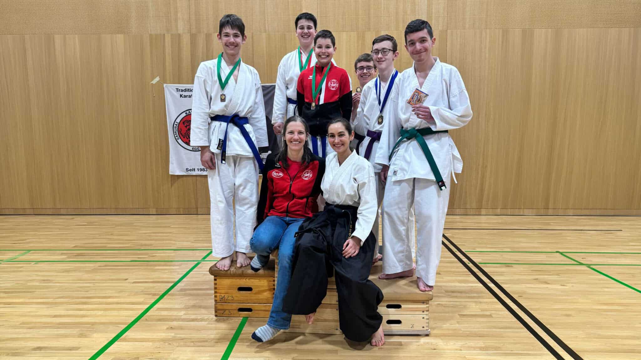 Gruppenfoto mit den Bludenzer Karatekas und ihren Betreuerinnen im Gi bzw. Hakama mit Medaillen in der Halle nach der Siegerehrung