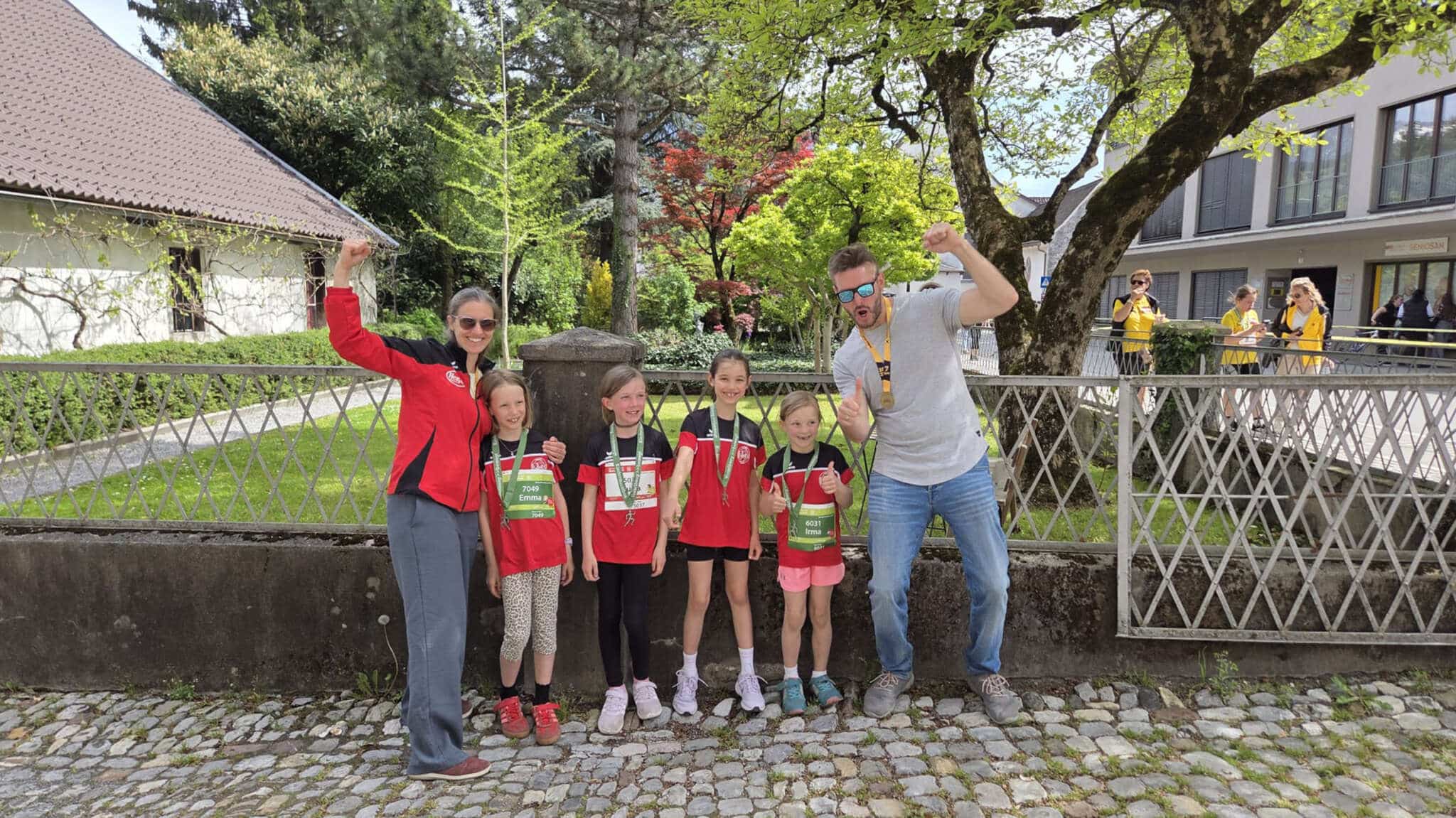 Gruppenfoto mit den vier Starterinnen des Kindermarathons und den zwei Trainern in Laufklamotten mit Medaillen nach dem Lauf auf der Straße vor einem Baum