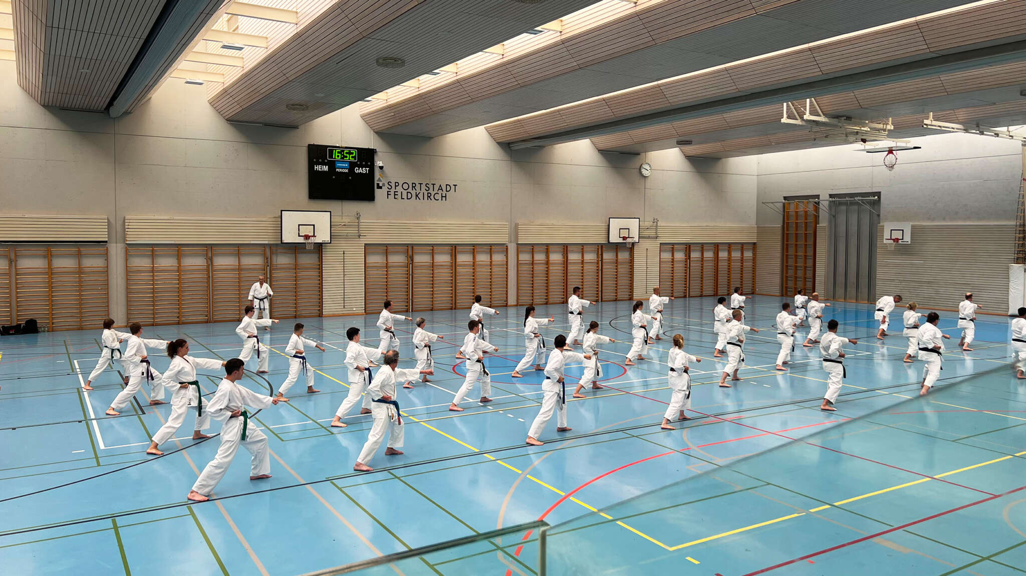 Eine Gruppe von mehreren Dutzend Karatekas führt Oi Zuki Chudan unter den Augen von Meister Fugazza aus. Die Karatekas blicken nach rechts und sind auf mehrere Reihen aufgeteilt. Sie stehen in einer Halle in der Stellung Zenkutsu Dachi.